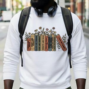 May include: White sweatshirt with a graphic of books and flowers. The book spines have phrases like "Dream Like Martin" and "Challenge Like Rosa." The design uses brown, orange, and green. A stylish and educational garment.