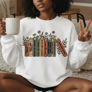 May include: White sweatshirt with a graphic of books and flowers. The books are inscribed with phrases such as "Dream Like Martin" and "Challenge Like Rosa." A relaxed, comfortable garment.