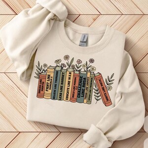 May include: A cream-coloured sweatshirt with a graphic of books, each with a motivational title such as "Fight Like Malcolm X" and "Educate Like W.E.B." Floral and leaf designs are between the books. The sweatshirt has a crew neck.