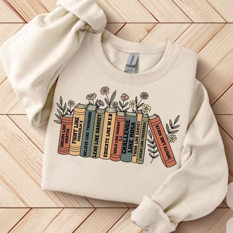 May include: A cream-coloured sweatshirt with a graphic of books, each with a motivational title such as "Fight Like Malcolm X" and "Educate Like W.E.B." Floral and leaf designs are between the books. The sweatshirt has a crew neck.