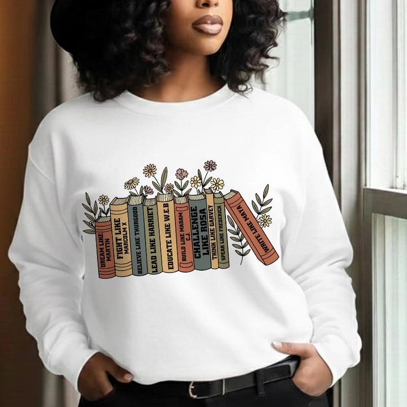 May include: White sweatshirt with a graphic of books in various colours, each inscribed with motivational quotes such as "Dream Like Martin" and "Write Like Maya." The books are decorated with floral and leaf motifs.