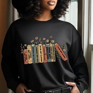 May include: Black sweatshirt with a graphic of books, including titles such as "Fight Like Malcolm X" and "Write Like Maya." The design incorporates floral elements. A casual, comfortable top.