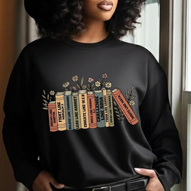 May include: Black sweatshirt with a graphic of books, including titles such as "Fight Like Malcolm X" and "Write Like Maya." The design incorporates floral elements. A casual, comfortable top.