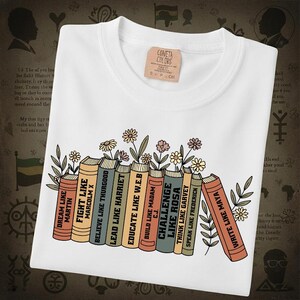 May include: White t-shirt with a graphic of books and flowers. The books have titles like "Dream Like Martin" and "Write Like Maya". A small tag on the shirt reads "CONETA COLORS".