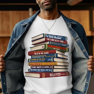 May include: White t-shirt featuring a stack of books with titles like "Beloved" and "The Fire Next Time." The books are in various colors, including red, blue, and green. The shirt is worn by a person wearing a denim jacket.