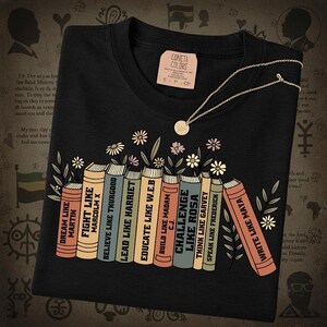 May include: Black t-shirt featuring a graphic of books with titles such as "Fight Like Malcolm X" and "Challenge Like Rosa." A gold necklace with a small pendant is on the shirt. The tag on the shirt says "Cometa Colors."