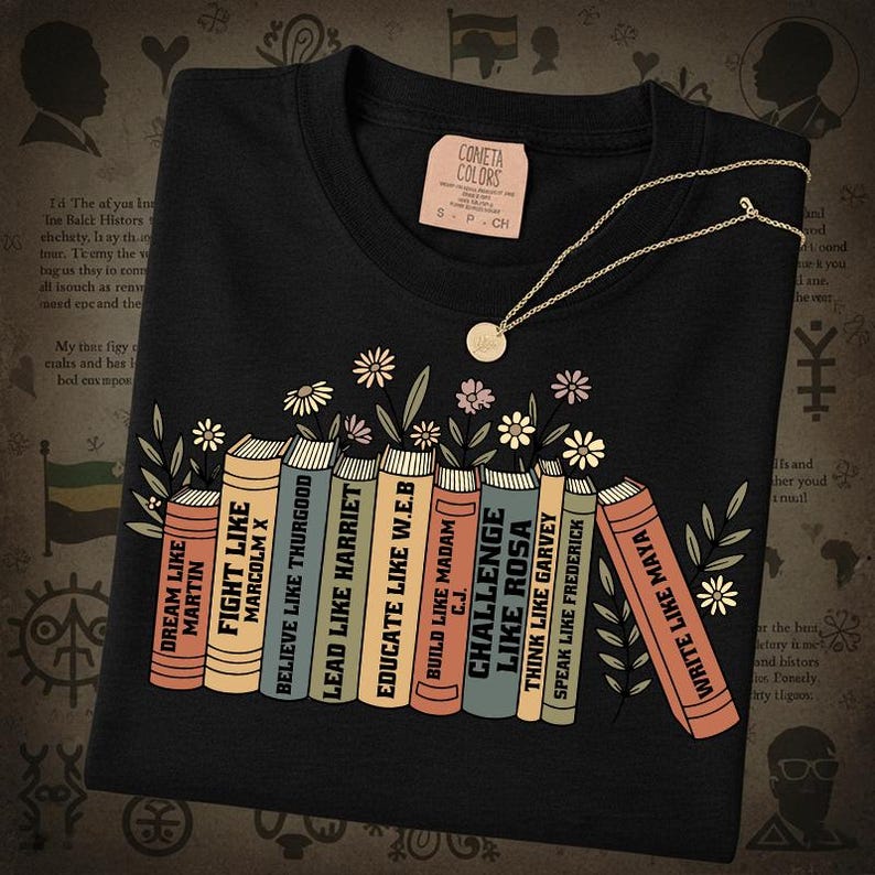 May include: Black t-shirt featuring a graphic of books with titles such as "Fight Like Malcolm X" and "Challenge Like Rosa." A gold necklace with a small pendant is on the shirt. The tag on the shirt says "Cometa Colors."
