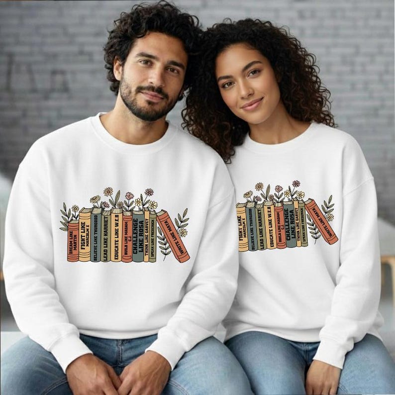 May include: White sweatshirts featuring a graphic of books and flowers. The books display phrases such as "Dream Like Martin" and "Challenge Like Rosa." The sweatshirts are worn by a man and a woman.