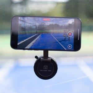 May include: A smartphone mounted on a black holder, displaying a live video of a blue sports court. The phone is recording a sporting event. The holder has the text "THE PADEL AXIS" printed on it.