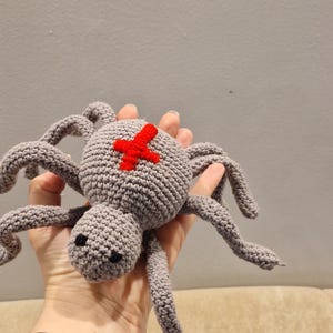 May include: A crocheted spider toy in shades of gray, featuring eight long, curved legs and a round body. A red cross is stitched on the spider's back. The toy is held in a hand.
