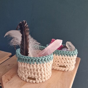 May include: Two small, cream-colored crochet baskets with a teal trim, filled with feathers and crystals. The baskets sit on a wooden surface against a teal background. One basket contains a large, clear quartz crystal.