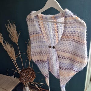 May include: A hand-knitted shawl in a soft, textured yarn, featuring a mix of white, lavender, and peach hues. The shawl is fastened with two decorative buttons and hangs on a white hanger. The overall style is cozy and handmade.
