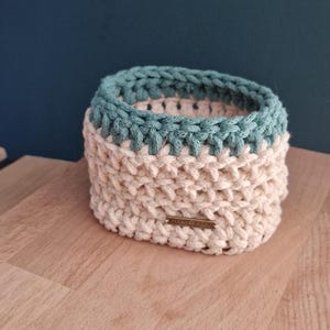 May include: A handmade, square crochet basket with a two-tone design. The bottom half is a cream color, and the top rim is a teal color. A small, rectangular tag is attached to the front. The basket is sitting on a wooden surface.