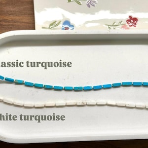 May include: Two strands of turquoise beads, one classic blue and the other white, are displayed on a white tray. The text "Classic turquoise" and "White turquoise" are above and below the strands, respectively. The background includes a floral design.