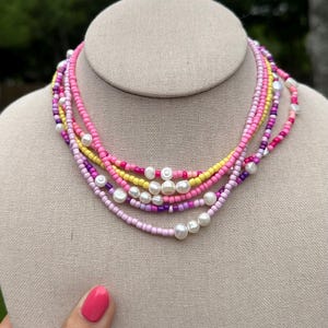 May include: A multi-strand beaded necklace featuring layers of small glass seed beads in shades of pink, purple, and yellow, interspersed with irregular white freshwater pearls. The necklace consists of several distinct strands worn together.