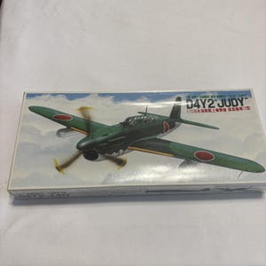 May include: A model kit box featuring a green and red Japanese Navy carrier dive-bomber plane, the D4Y2 "Judy". The plane has a propeller and red circle markings on the wings. The box has text in English and Japanese.