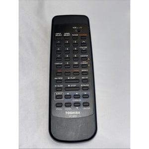 May include: Black Toshiba VC-653 VCR remote control with numerous buttons for power, channel selection, and playback. The remote has a curved design and is labeled with various functions.