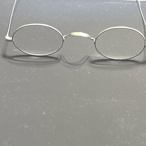 Könnte beinhalten: Silberfarbene, oval geformte Brille mit dünnen Rahmen und einem Steg. Die Gläser sind klar, und die Bügel erstrecken sich zu den Seiten. Die Brille ist für die Sehkorrektur bestimmt, möglicherweise Vintage oder antik.