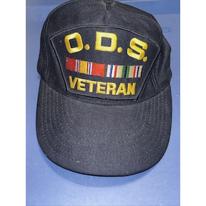 Puede incluir: Gorra de béisbol negra con visera curva. La gorra presenta un parche con el texto "O.D.S. VETERAN" en letras doradas, junto con emblemas de cintas de colores. La gorra está hecha de tela texturizada.