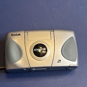 以下が含まれることがあります： グレーのアクセントが付いたシルバーのKodak Advantix C350カメラ。 カメラは、丸みを帯びたエッジとレンズカバーを備えた長方形の形状をしています。 Kodakのロゴが左上に表示されています。 青い表面にあります。
