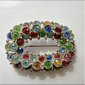 May include: Oval-shaped vintage brooch with a silver-tone metal frame. The brooch is adorned with a variety of colorful, round gemstones in shades of red, blue, green, yellow, and pink. The pin closure is visible on the back.