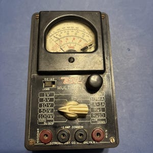 May include: Vintage black ZEOL multimeter with a round analog display and multiple settings. Features a selector dial, input jacks, and the text "ZEOL MULTIMETER". The device measures voltage, current, and resistance.