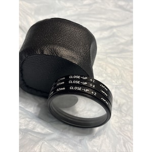 May include: A black leather camera lens case next to a set of three close-up camera lenses. The lenses are labeled with the text "CLOSE-UP +1", "CLOSE-UP +4", and "CLOSE-UP +2". The lenses are 52mm.