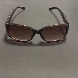 May include: Sunglasses with a tortoiseshell pattern frame and brown gradient lenses. The sunglasses have a classic oval shape and dark brown temples. The lenses are a medium tint, providing sun protection. The sunglasses are suitable for everyday wear.