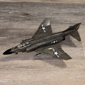 May include: A dark gray model fighter jet with a black nose cone and a clear cockpit canopy. The fuselage features the text U.S. AIR FORCE and white star insignias with red and white stripes on the wings and sides of the aircraft body.