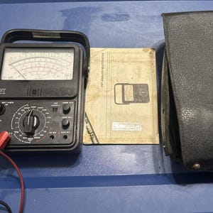 May include: A black FET analog multimeter with a handle, a dial, and multiple settings. A paper manual and a black leather case are next to the multimeter. The multimeter has red and black test leads.