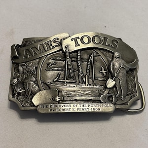 May include: A silver-colored metal belt buckle with the words "AMES TOOLS" at the top. The buckle features a detailed relief scene of a ship, a polar explorer, and the text "THE DISCOVERY OF THE NORTH POLE BY ROBERT E. PEARY-1909."