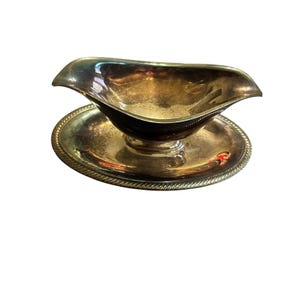 May include: Silver-plated gravy boat with a matching round base. The gravy boat has a curved spout and a decorative rim. The base has a rope-like edge. The set is designed for serving sauces and gravies.