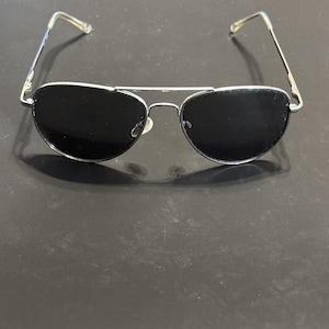 Puede incluir: Gafas de sol de aviador con montura de metal plateado y lentes oscuras. Las gafas tienen un doble puente y patillas de plástico transparente. Las lentes son de forma ovalada.