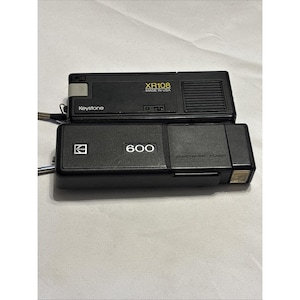 May include: Two black vintage cameras. The top camera is a Keystone XR108, made in the USA. The bottom camera is a Kodak 600 with electronic flash.