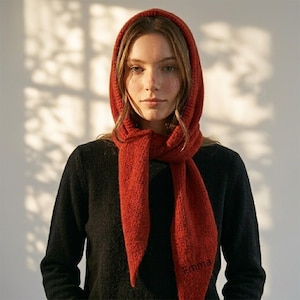 May include: A knitted scarf and hood in a rust colour. The scarf is wrapped around the neck and over the head, with the hood framing the face. The word "Emma" is embroidered on the scarf. The person is wearing a black jumper.
