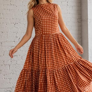 May include: A sleeveless maxi dress featuring a rust orange and white gingham check pattern. The garment has a high round neckline, a fitted bodice, and a voluminous, tiered skirt that flares out from the waistline to the floor in a full silhouette.
