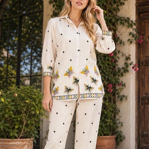 May include: A cream-coloured button-down shirt and matching trousers set featuring a repeating black diamond pattern and colourful embroidered butterfly motifs at the hem. The sleeves and pant cuffs are finished with a decorative geometric border.