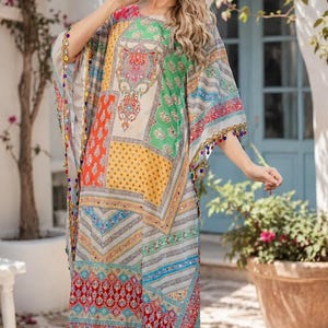 May include: A long, loose-fitting kaftan featuring a vibrant patchwork design with geometric and floral patterns in shades of green, orange, red, and blue. The garment is accented with small, dangling colorful beads along the hem and sleeves.