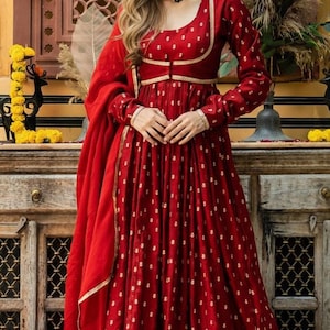 May include: A long-sleeved, floor-length red dress featuring a repeating gold floral pattern, a scoop neckline, and gold trim along the bodice. The dress is paired with a matching red dupatta draped over the shoulder, accented by a white beaded choker.