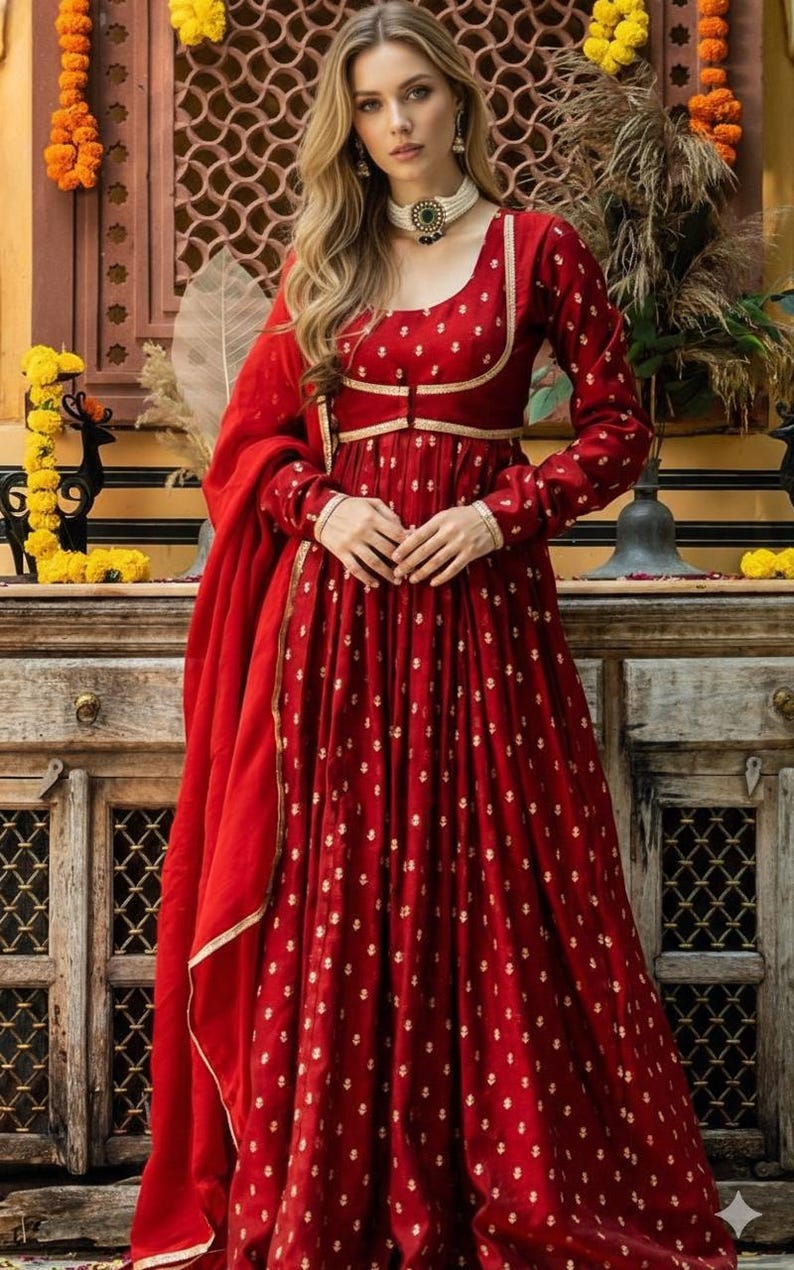 May include: A long-sleeved, floor-length red dress featuring a repeating gold floral pattern, a scoop neckline, and gold trim along the bodice. The dress is paired with a matching red dupatta draped over the shoulder, accented by a white beaded choker.