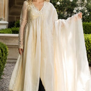 May include: A cream-colored long-sleeved tunic with a V-neckline featuring silver mirror embellishments, paired with a matching sheer dupatta and dark trousers. The tunic has a flowing silhouette with a central front slit and sheer sleeves.