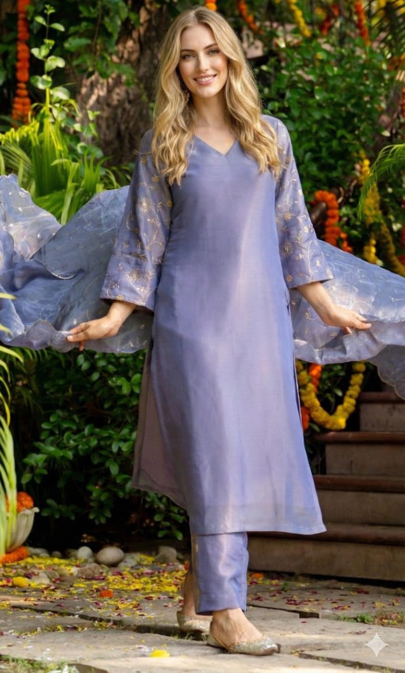 May include: A periwinkle-colored, two-piece outfit. The top has a V-neck and three-quarter sleeves with gold embroidery. The matching pants complete the ensemble. A sheer, flowing scarf adds movement. The outfit is worn outdoors.