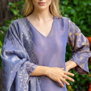 May include: A periwinkle-colored, long-sleeved dress with a V-neck and matching shawl. The dress and shawl feature gold floral embroidery along the sleeves and edges. The outfit is made of a shiny, flowing fabric.
