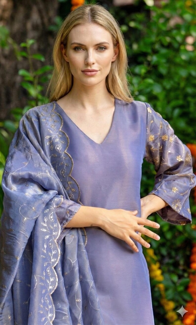 May include: A periwinkle-colored, long-sleeved dress with a V-neck and matching shawl. The dress and shawl feature gold floral embroidery along the sleeves and edges. The outfit is made of a shiny, flowing fabric.