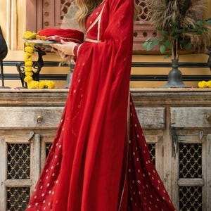 May include: A vibrant red floor-length dress featuring a subtle gold-toned pattern, a matching red dupatta draped over the shoulder, and a gold-trimmed neckline. The garment is paired with a white choker necklace and gold-toned wrist bangles.