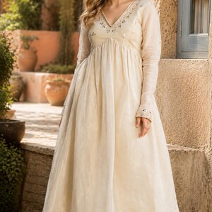 May include: A cream-colored long-sleeved dress with a V-neckline featuring intricate embroidery and small mirror embellishments. The dress has a fitted bodice that transitions into a flowing, pleated skirt, paired with matching wide-leg trousers.