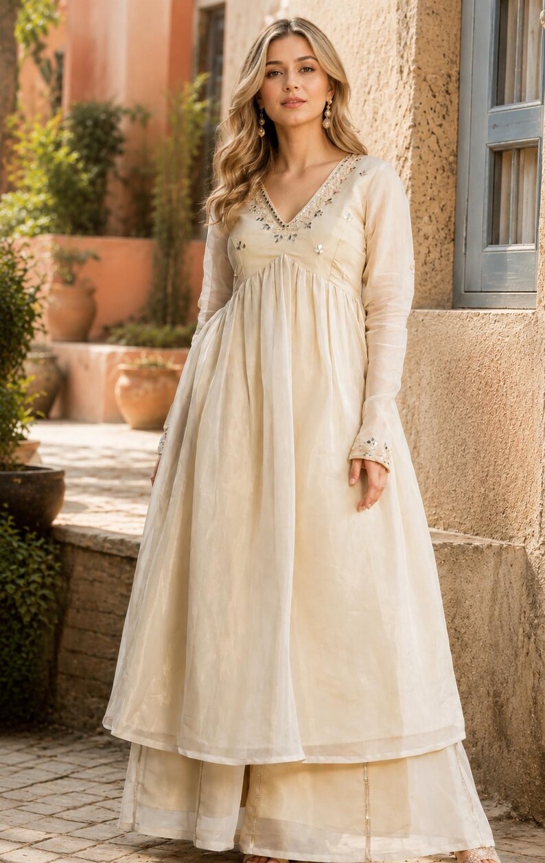 May include: A cream-colored long-sleeved dress with a V-neckline featuring intricate embroidery and small mirror embellishments. The dress has a fitted bodice that transitions into a flowing, pleated skirt, paired with matching wide-leg trousers.