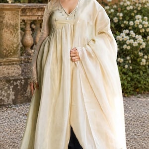 May include: A cream-colored long-sleeved tunic with a V-neckline featuring silver embroidery and mirror accents, paired with a matching sheer dupatta and a solid black skirt. The tunic has a gathered waist and a flowing, layered silhouette.