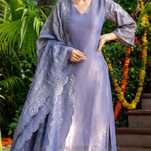 May include: A periwinkle-colored salwar kameez ensemble. The outfit includes a V-neck tunic, matching pants, and a dupatta with a scalloped edge. The sleeves and dupatta feature gold-colored embellishments. The outfit is worn with gold-colored flats.