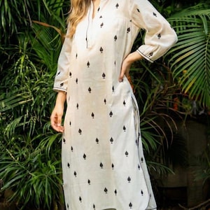 Può includere: Completo tunica e pantaloni color crema, ornato da motivi neri a forma di picche. La tunica a maniche lunghe ha un colletto alla coreana e bordi neri. I pantaloni sono a gamba dritta. L'outfit è abbinato a ballerine dorate. Sfondo di fogliame verde.